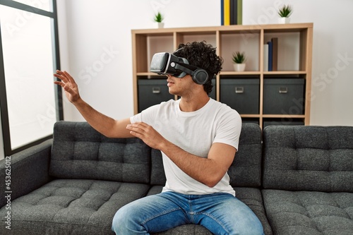 Young hispanic man playing with virtual reality glasses sitting on the sofa at home. photo