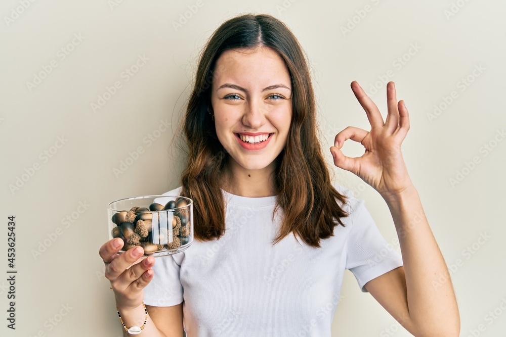 Young brunette woman holding raw hazelnuts doing ok sign with fingers, smiling friendly gesturing excellent symbol
