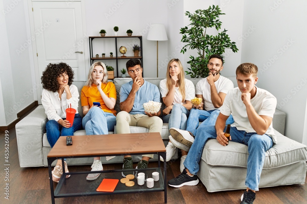 Group of people eating snack sitting on the sofa at home serious face thinking about question with hand on chin, thoughtful about confusing idea