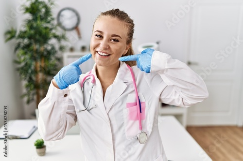 Young caucasian woman wearing doctor uniform and stethoscope at the clinic smiling cheerful showing and pointing with fingers teeth and mouth. dental health concept.