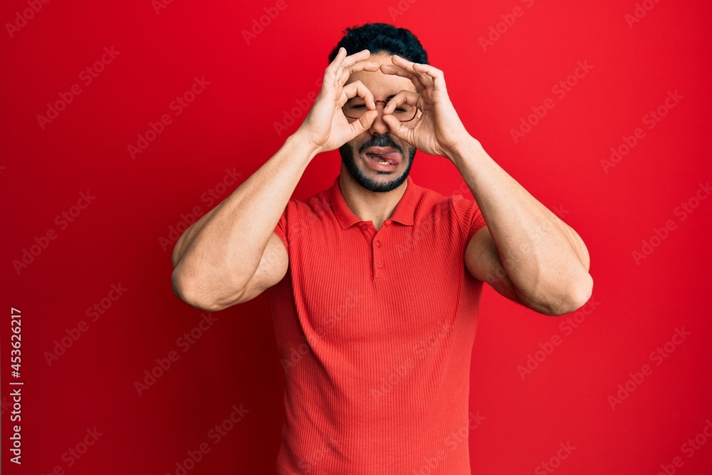 Young hispanic man wearing casual clothes and glasses doing ok gesture like binoculars sticking tongue out, eyes looking through fingers. crazy expression. Young hispanic man wearing casual clothes and glasses doing ok gesture like binoculars sticking tongue out, eyes looking through fingers. crazy expression.