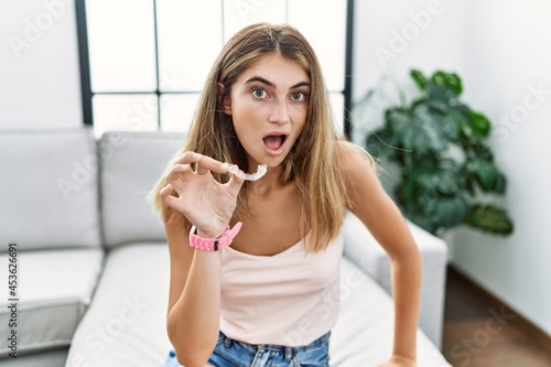 Young blonde woman holding invisible aligner orthodontic in shock face  looking skeptical and sarcastic  surprised with open mouth