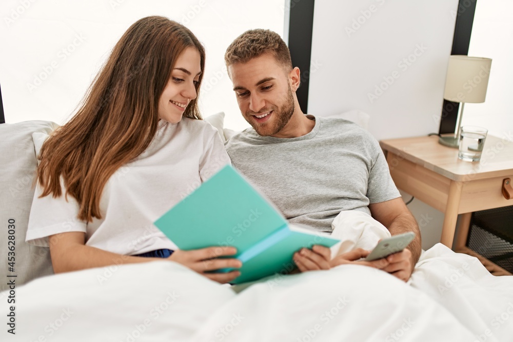 Young caucasian couple reading book and using smartphone at bed. Young caucasian couple reading book and using smartphone at bed.