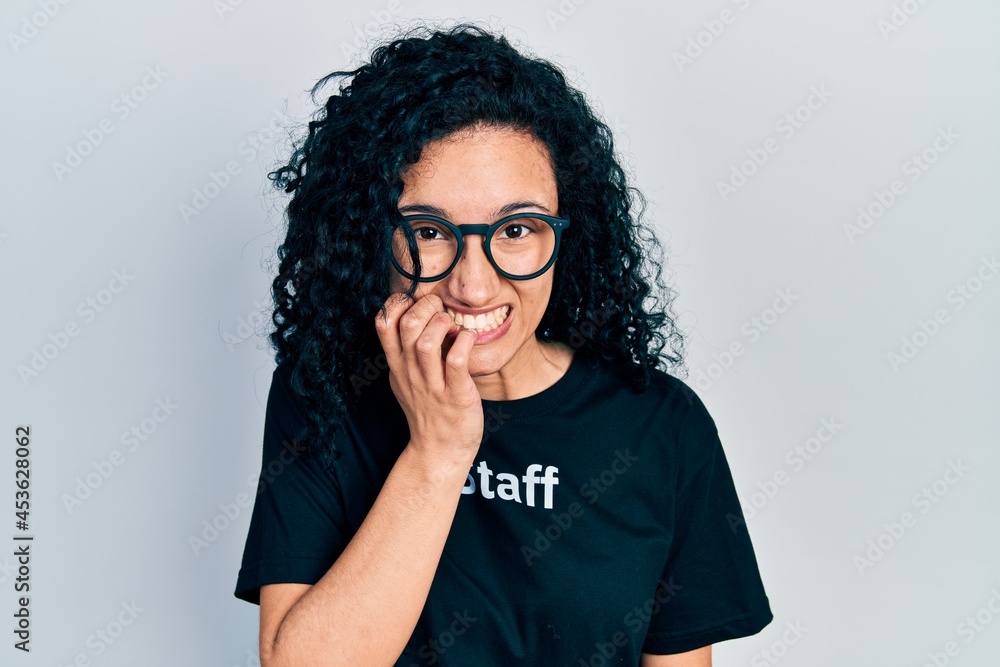 Young hispanic woman with curly hair wearing staff t shirt looking stressed and nervous with hands on mouth biting nails. anxiety problem. Young hispanic woman with curly hair wearing staff t shirt looking stressed and nervous with hands on mouth biting nails. anxiety problem.