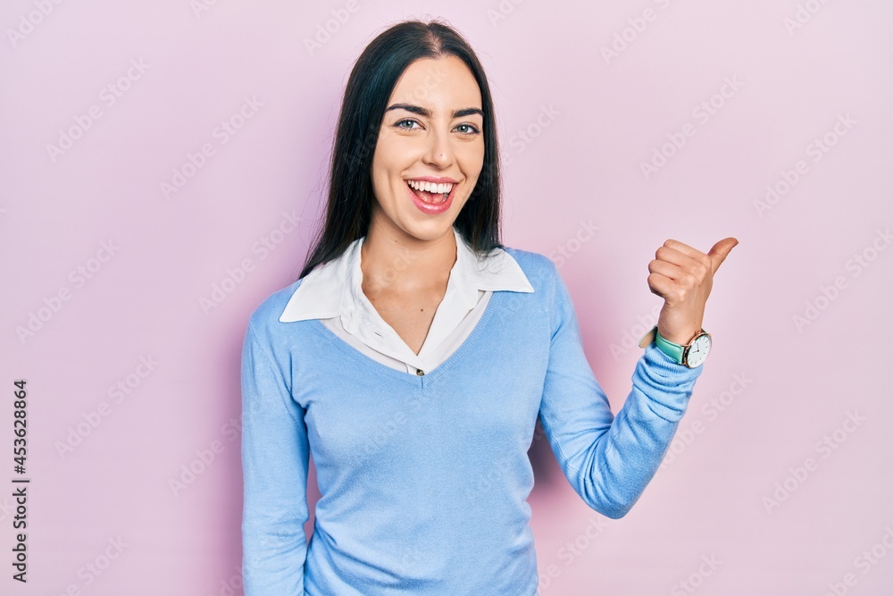 Beautiful woman with blue eyes standing over pink background smiling with happy face looking and pointing to the side with thumb up. Beautiful woman with blue eyes standing over pink background smiling with happy face looking and pointing to the side with thumb up.