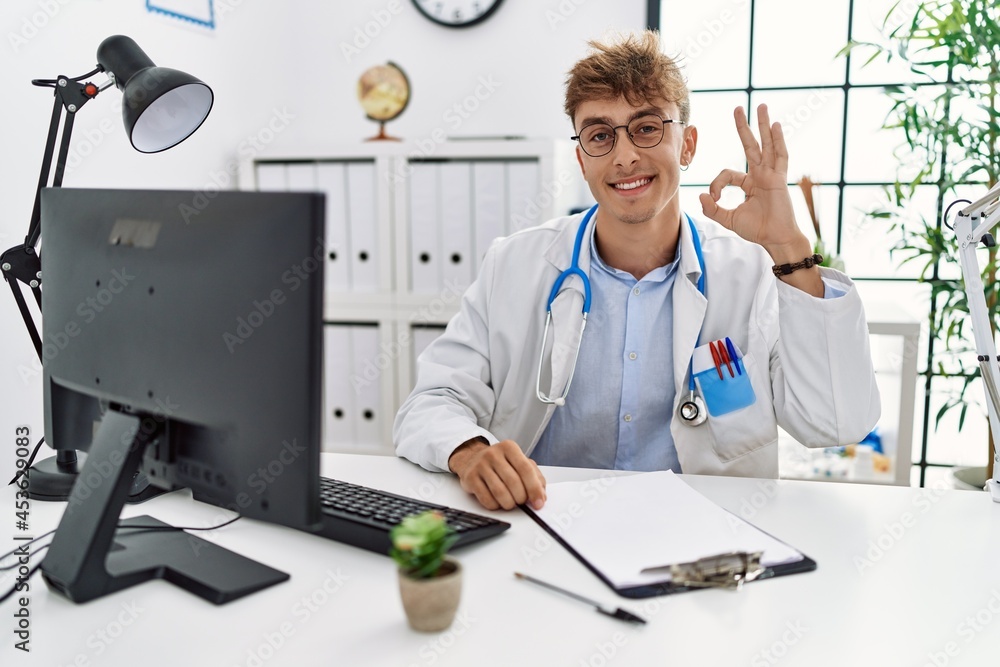 Young caucasian doctor man working at the clinic smiling positive doing ok sign with hand and fingers. successful expression. Young caucasian doctor man working at the clinic smiling positive doing ok sign with hand and fingers. successful expression.