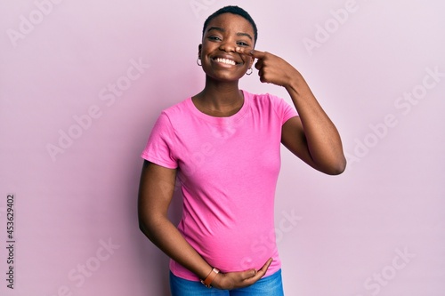 Young african american woman expecting a baby  touching pregnant belly pointing with hand finger to face and nose  smiling cheerful. beauty concept