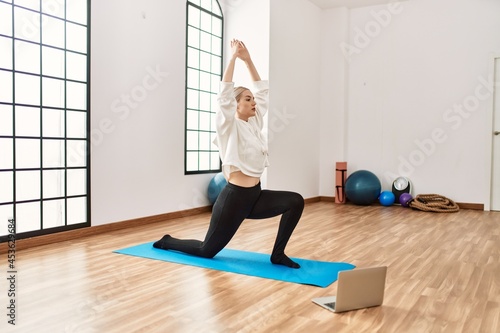 Young blonde girl concentrated having online yoga class using laptop at sport center.