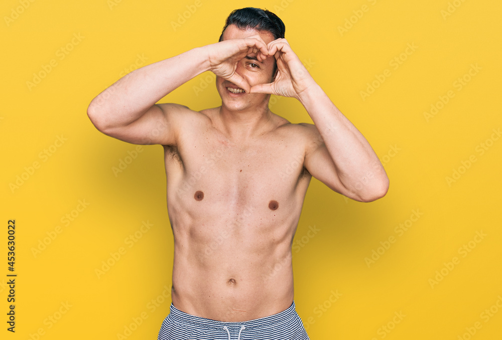 Handsome young man wearing swimwear shirtless doing heart shape with hand and fingers smiling looking through sign