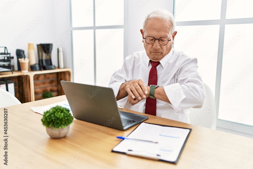Senior man working at the office using computer laptop checking the time on wrist watch, relaxed and confident Senior man working at the office using computer laptop checking the time on wrist watch, relaxed and confident