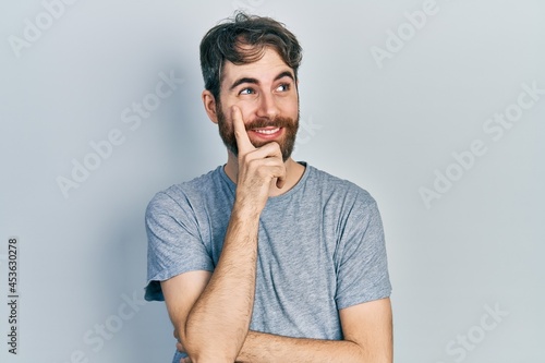 Caucasian man with beard wearing casual grey t shirt with hand on chin thinking about question, pensive expression. smiling with thoughtful face. doubt concept.