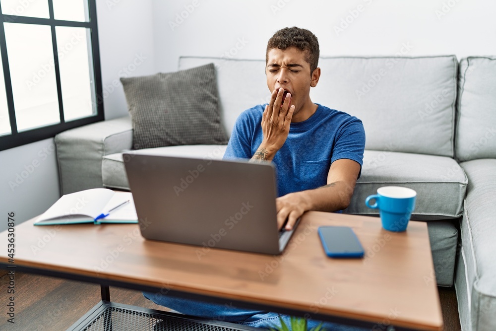 Young handsome hispanic man using laptop sitting on the floor bored yawning tired covering mouth with hand. restless and sleepiness. Young handsome hispanic man using laptop sitting on the floor bored yawning tired covering mouth with hand. restless and sleepiness.