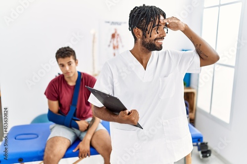 Young hispanic man working at pain recovery clinic with a man with broken arm very happy and smiling looking far away with hand over head. searching concept.