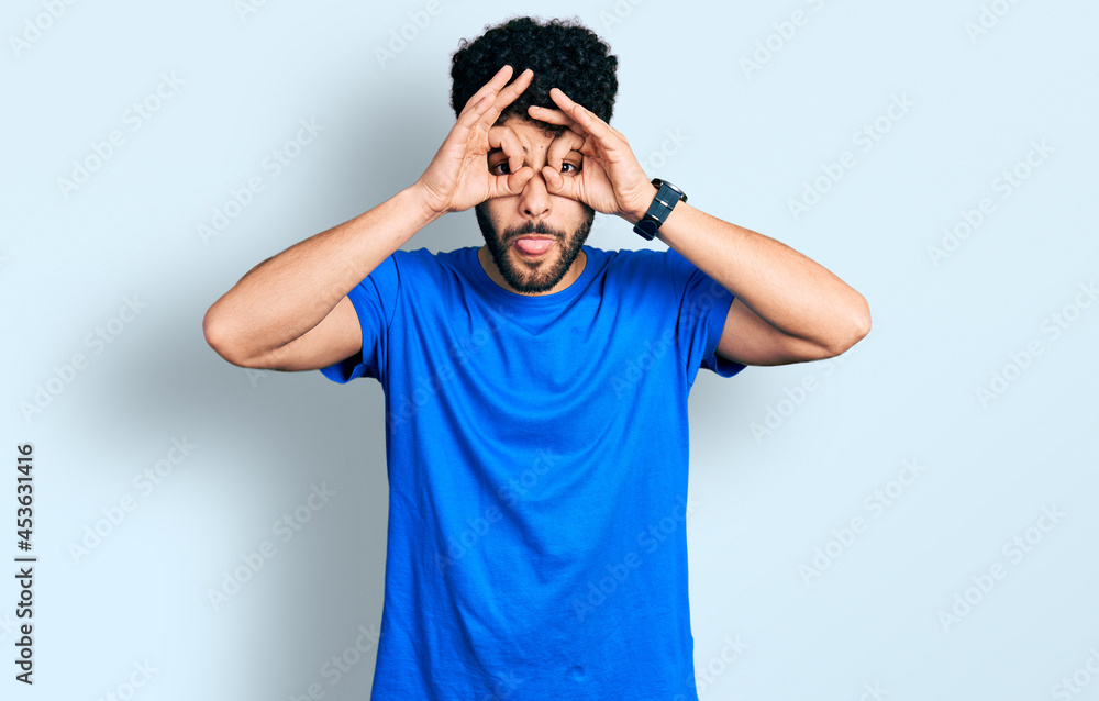 Young arab man with beard wearing casual blue t shirt doing ok gesture like binoculars sticking tongue out, eyes looking through fingers. crazy expression. Young arab man with beard wearing casual blue t shirt doing ok gesture like binoculars sticking tongue out, eyes looking through fingers. crazy expression.