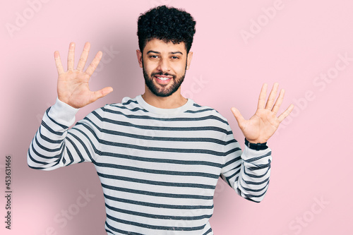 Young arab man with beard wearing casual striped sweater showing and pointing up with fingers number ten while smiling confident and happy.