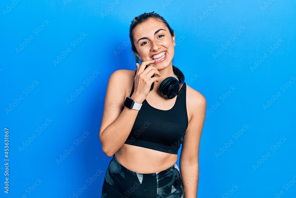 Beautiful hispanic woman wearing gym clothes and using headphones looking confident at the camera with smile with crossed arms and hand raised on chin. thinking positive. Beautiful hispanic woman wearing gym clothes and using headphones looking confident at the camera with smile with crossed arms and hand raised on chin. thinking positive.
