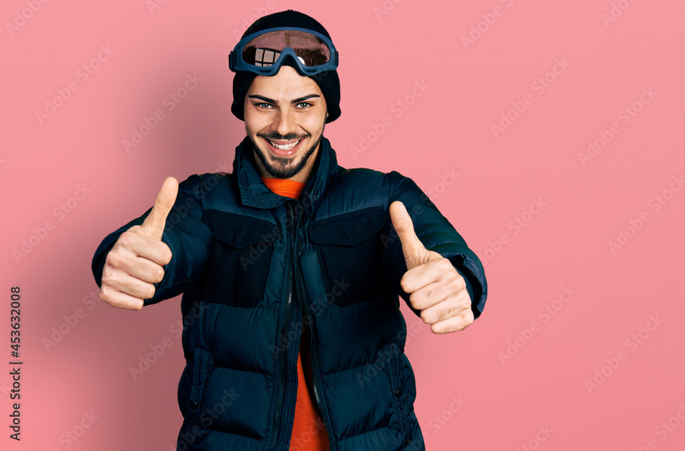 Young hispanic man with beard wearing snow wear and sky glasses approving doing positive gesture with hand, thumbs up smiling and happy for success. winner gesture. Young hispanic man with beard wearing snow wear and sky glasses approving doing positive gesture with hand, thumbs up smiling and happy for success. winner gesture.