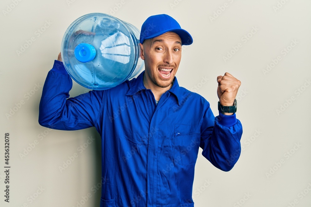 Bald courier man with beard holding a gallon bottle of water for delivery screaming proud, celebrating victory and success very excited with raised arms