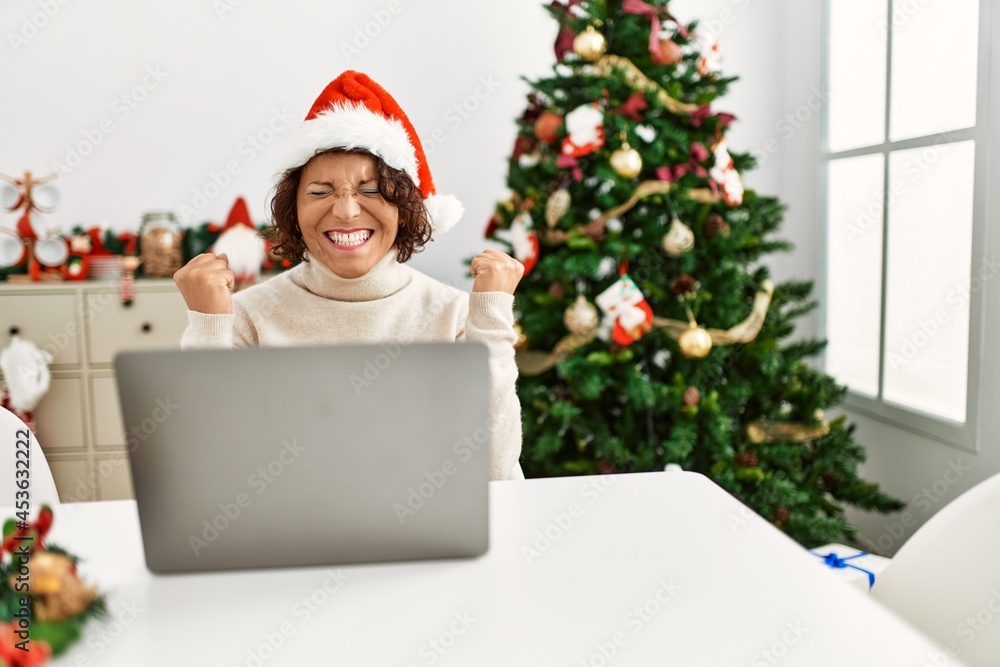 Middle age hispanic woman using laptop sitting by christmas tree very happy and excited doing winner gesture with arms raised, smiling and screaming for success. celebration concept. Middle age hispanic woman using laptop sitting by christmas tree very happy and excited doing winner gesture with arms raised, smiling and screaming for success. celebration concept.