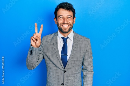 Handsome man with beard wearing business suit and tie showing and pointing up with fingers number two while smiling confident and happy.