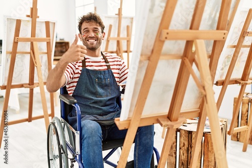 Young handsome man with beard at art studio sitting on wheelchair smiling happy and positive, thumb up doing excellent and approval sign
