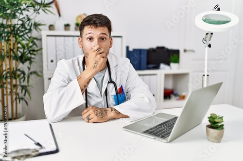 Young doctor working at the clinic using computer laptop smelling something stinky and disgusting, intolerable smell, holding breath with fingers on nose. bad smell