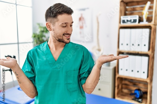 Young physiotherapist man working at pain recovery clinic smiling showing both hands open palms  presenting and advertising comparison and balance
