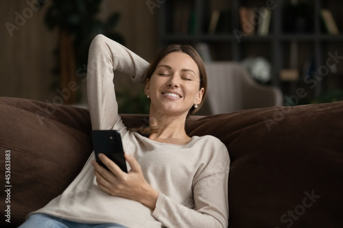 Smiling distracted woman with closed eyes holding smartphone, relaxing on cozy couch at home, pleasant attractive young female enjoying lazy leisure time with device, chatting in social networks