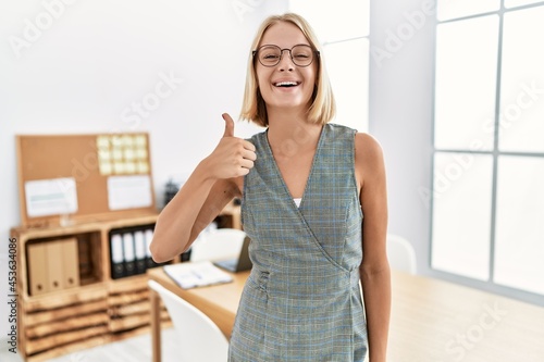 Young caucasian woman working at the office wearing glasses smiling happy and positive, thumb up doing excellent and approval sign