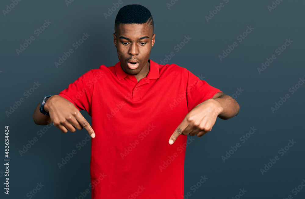 Fototapeta premium Young african american man wearing casual red t shirt pointing down with fingers showing advertisement, surprised face and open mouth