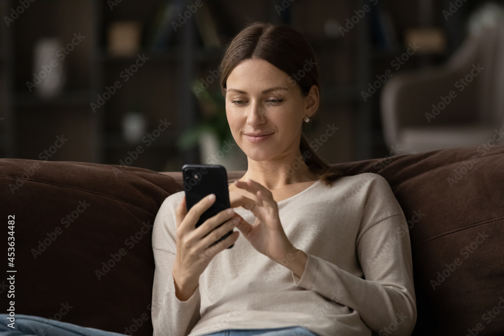 Close up satisfied young woman using smartphone, sitting on couch at home, pleasant young female looking at phone screen, typing writing message in social network, browsing mobile device apps Close up satisfied young woman using smartphone, sitting on couch at home, pleasant young female looking at phone screen, typing writing message in social network, browsing mobile device apps