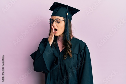 Young hispanic woman wearing graduation cap and ceremony robe bored yawning tired covering mouth with hand. restless and sleepiness.