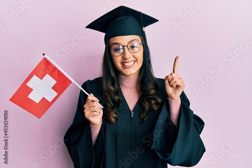 Young hispanic woman wearing graduation uniform holding switzerland flag smiling with an idea or question pointing finger with happy face, number one