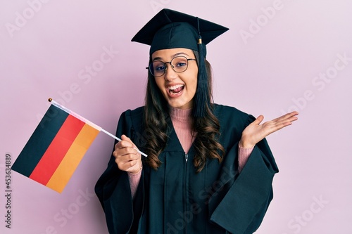 Young hispanic woman wearing graduation uniform holding germany flag celebrating achievement with happy smile and winner expression with raised hand
