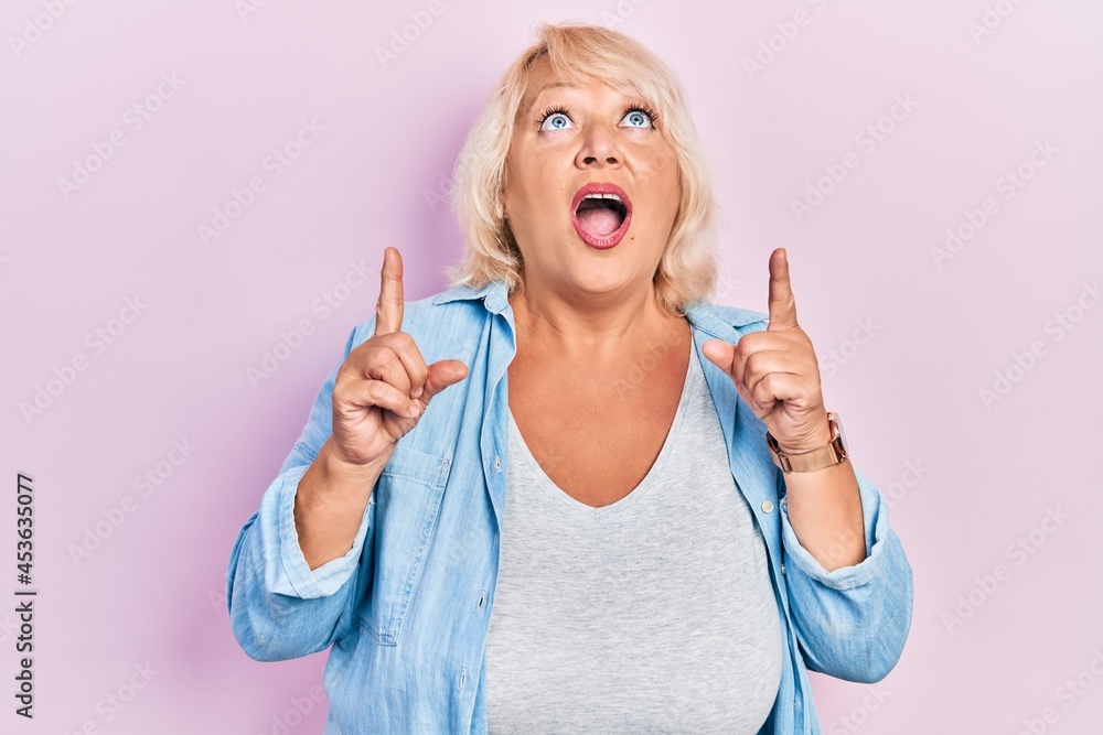 Middle age blonde woman wearing casual clothes amazed and surprised looking up and pointing with fingers and raised arms. Middle age blonde woman wearing casual clothes amazed and surprised looking up and pointing with fingers and raised arms.