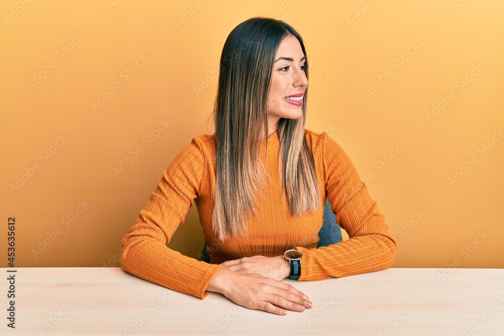 Young hispanic woman wearing casual clothes sitting on the table looking away to side with smile on face, natural expression. laughing confident. Young hispanic woman wearing casual clothes sitting on the table looking away to side with smile on face, natural expression. laughing confident.