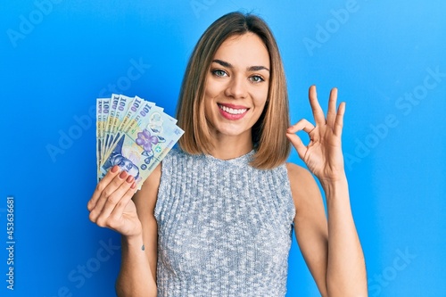 Young caucasian blonde woman holding 100 romanian leu banknotes doing ok sign with fingers, smiling friendly gesturing excellent symbol photo