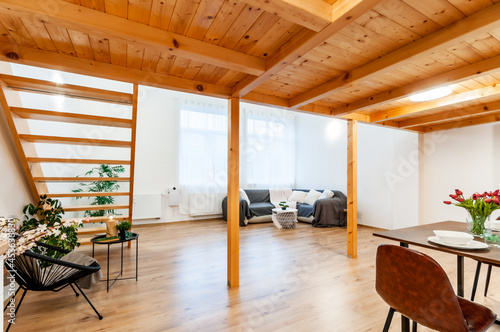 Bright loft apartment with large window, wooden floor and built-in staircase. Room has decorative plants, big corner sofa with few pillows and nice modern chair with small coffee table and small lamp.