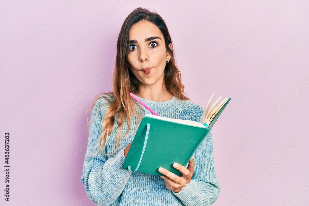 Young hispanic girl reading and writing book making fish face with mouth and squinting eyes, crazy and comical. Young hispanic girl reading and writing book making fish face with mouth and squinting eyes, crazy and comical.