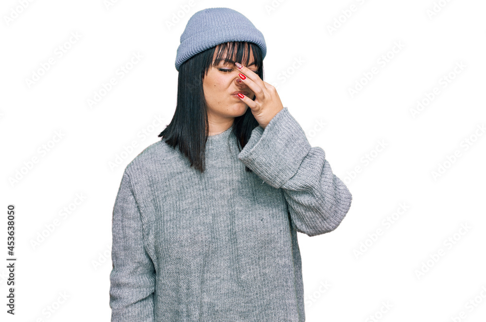 Young hispanic woman wearing cute wool cap smelling something stinky and disgusting, intolerable smell, holding breath with fingers on nose. bad smell