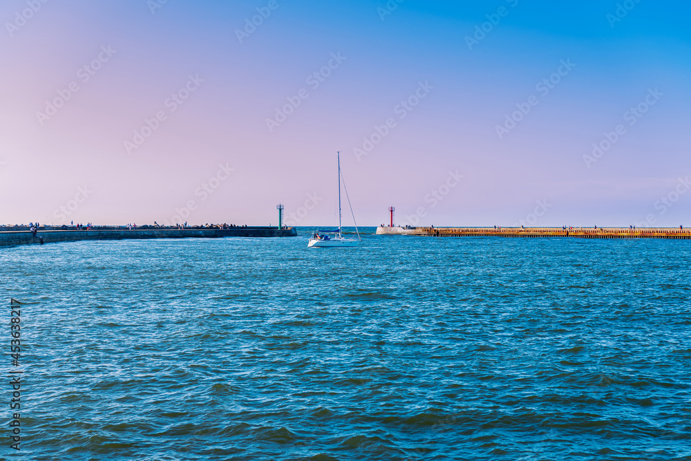 view of the bay in the port, a sailboat entering the port on a sunny day, concrete breakwaters in the background