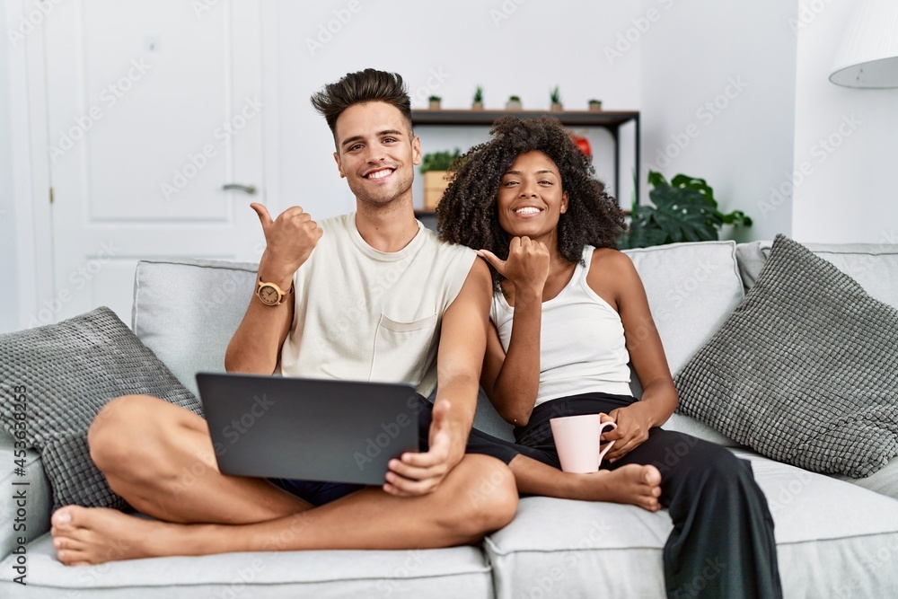 Young interracial couple using laptop at home sitting on the sofa pointing to the back behind with hand and thumbs up, smiling confident Young interracial couple using laptop at home sitting on the sofa pointing to the back behind with hand and thumbs up, smiling confident