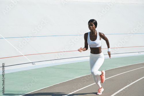 Confident young African woman in sports clothing running on track outdoors © gstockstudio