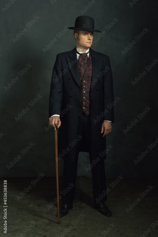 Young man in stylish vintage victorian attire. Standing with a cane in a room in front of a dark gray wall.
