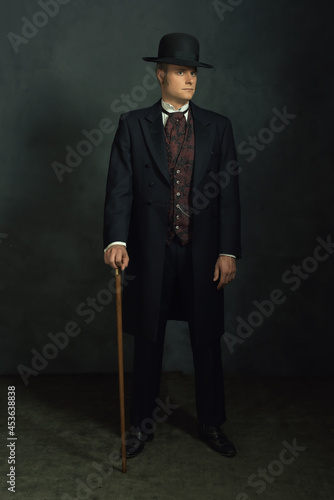 Obraz na płótnie Young man in stylish vintage victorian attire. Standing with a cane in a room in front of a dark gray wall. w salonie