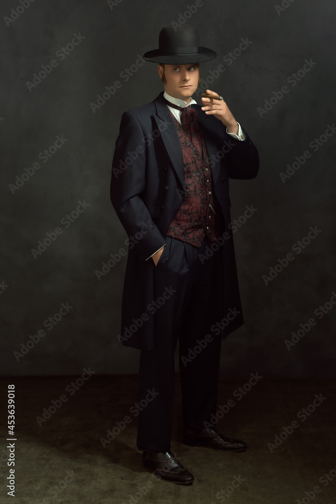 Cigar smoking young man in stylish vintage Victorian clothes stands in a room in front of a dark gray wall.