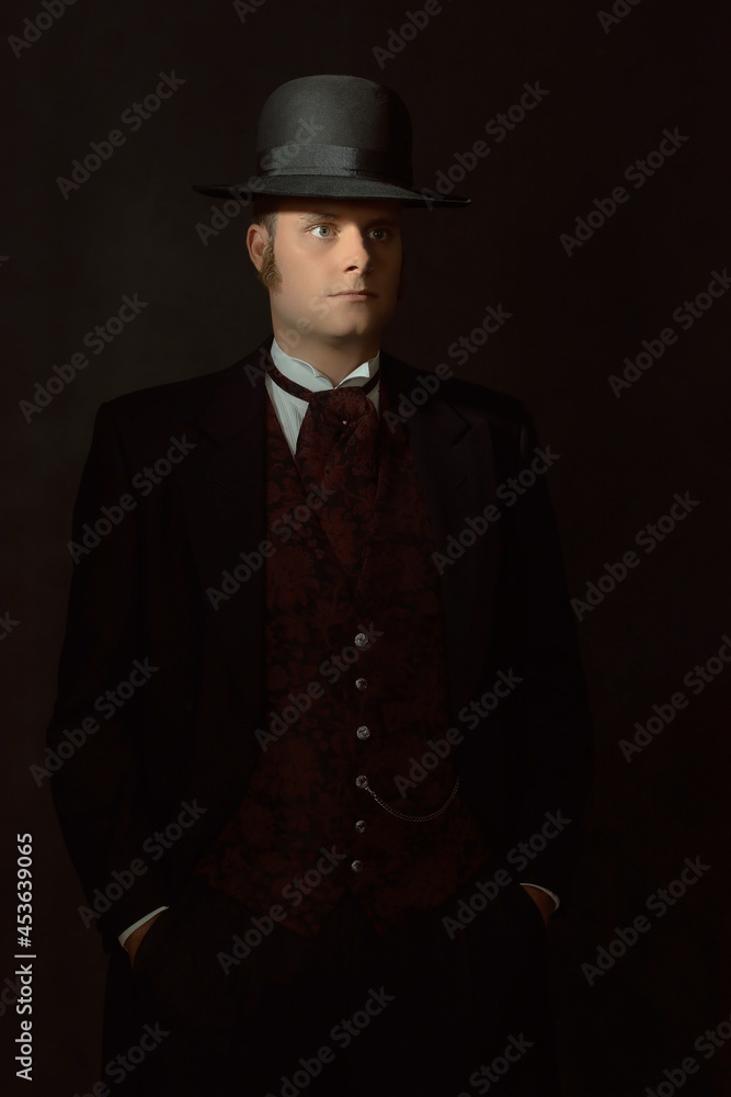 Shadowy portrait of a young man in stylish vintage Victorian attire in front of a dark gray wall.