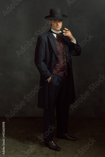 Obraz na płótnie Cigar smoking young man in stylish vintage Victorian clothes stands in a room in front of a dark gray wall. w salonie