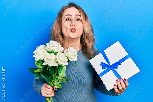 Middle age caucasian woman holding anniversary present and bouquet of flowers looking at the camera blowing a kiss being lovely and sexy. love expression.
