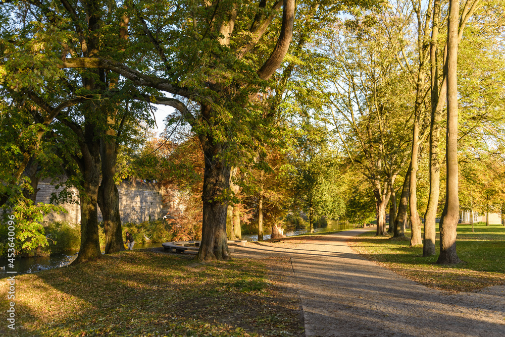 Outdoor scenery of walking way along Jeker canal and historical city wall at Monseigneur Nolenspark, city public park, in Autumn season, in Maastricht, Netherlands during evening sunset time.
